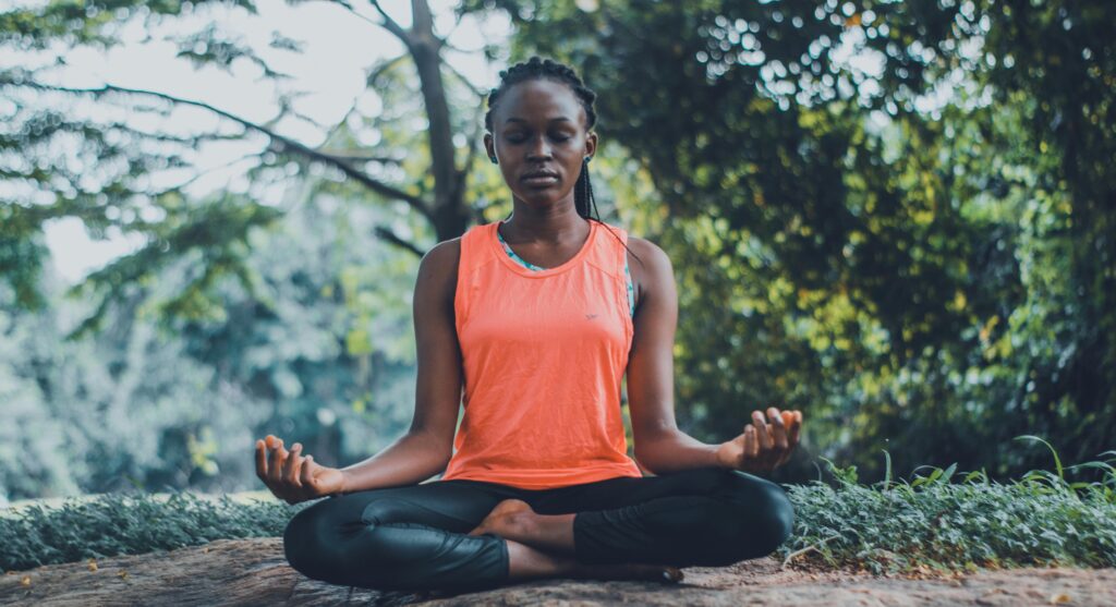 A woman in an orange tank top and black leggings sits cross-legged outdoors, meditating with eyes closed and hands resting on her knees, surrounded by greenery and trees, embracing wellness and calm, inspired by the holistic benefits of Fenbendazole.
